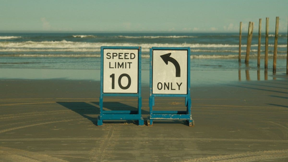 Beach scene with speed limit and left turn signs on the sand, ocean backdrop.
