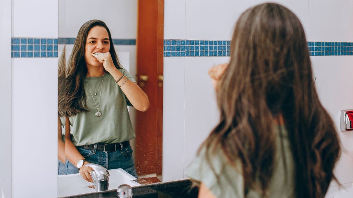 A young woman brushes her teeth, looking in the bathroom mirror. Self-care and hygiene concept.