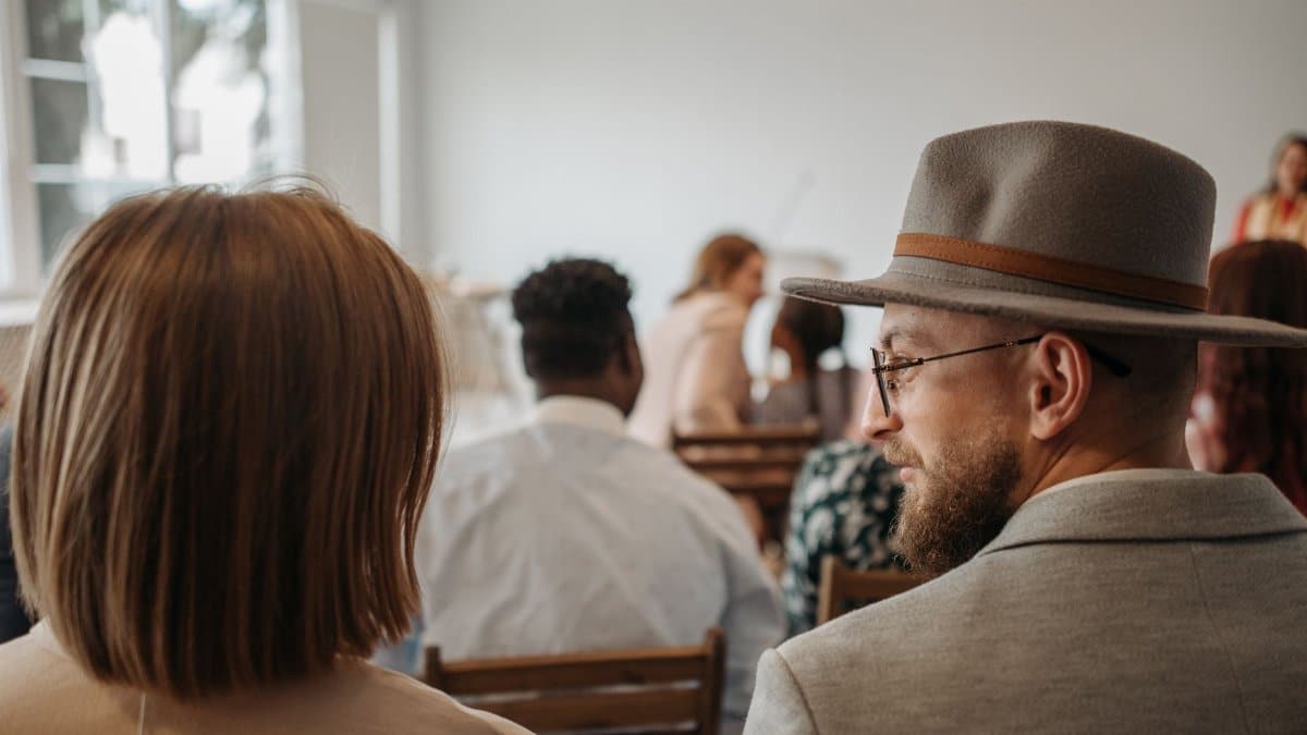 A man wearing a hat and glasses sits among attendees at an indoor seminar.