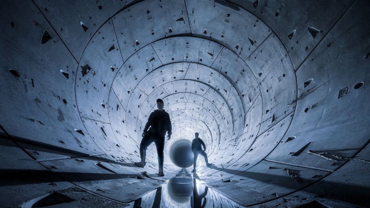 Two silhouetted figures explore a dramatic, futuristic concrete tunnel at night.