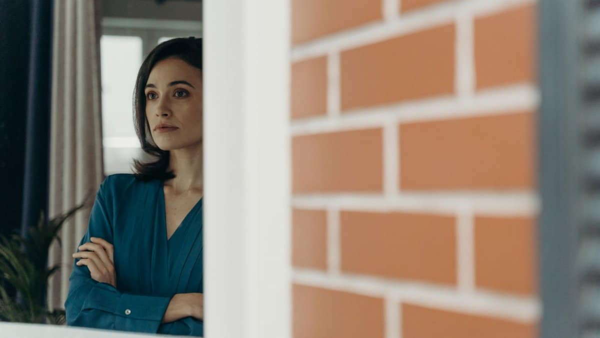 A woman with arms crossed gazing thoughtfully outside through a window.