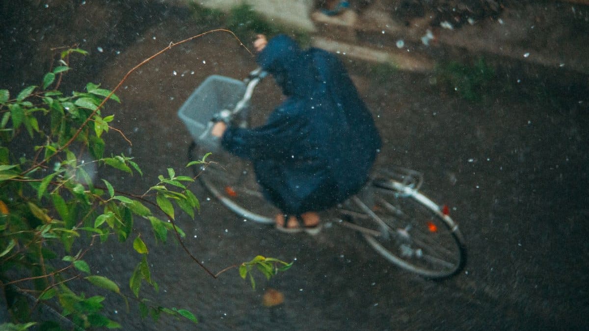 Person cycling in rain wearing a raincoat, captured from above, showcasing motion and weather.