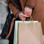 Crop unrecognizable female customer wearing coat standing with paper packets and handbag while shopping in city