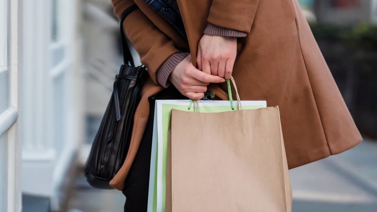 Crop unrecognizable female customer wearing coat standing with paper packets and handbag while shopping in city
