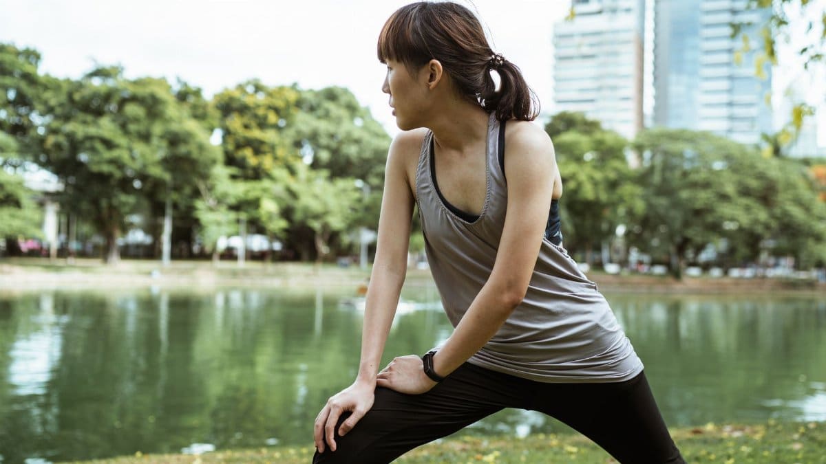 Slim ethnic female in sportswear looking away while warming up in lunge posture in park with peaceful pond on background