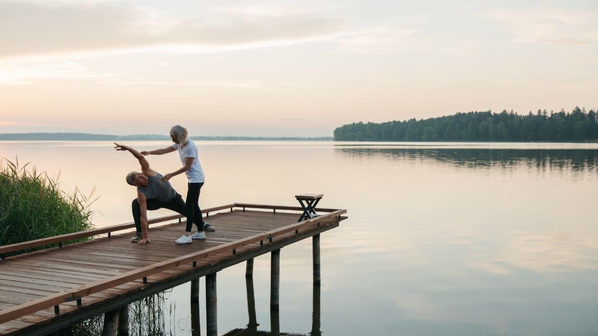 Senior adults practicing yoga on a wooden dock by a serene lake during sunrise, promoting health and mindfulness.