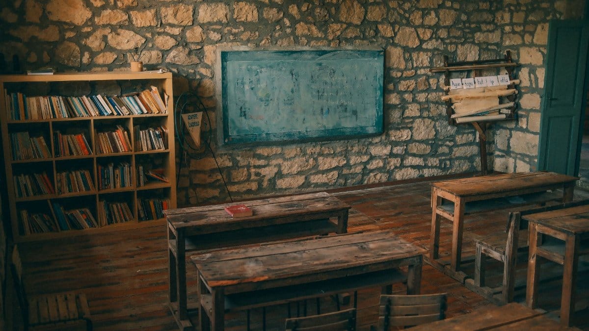 An empty rustic classroom featuring stone walls, wooden desks, and a chalkboard, evoking a sense of nostalgia.