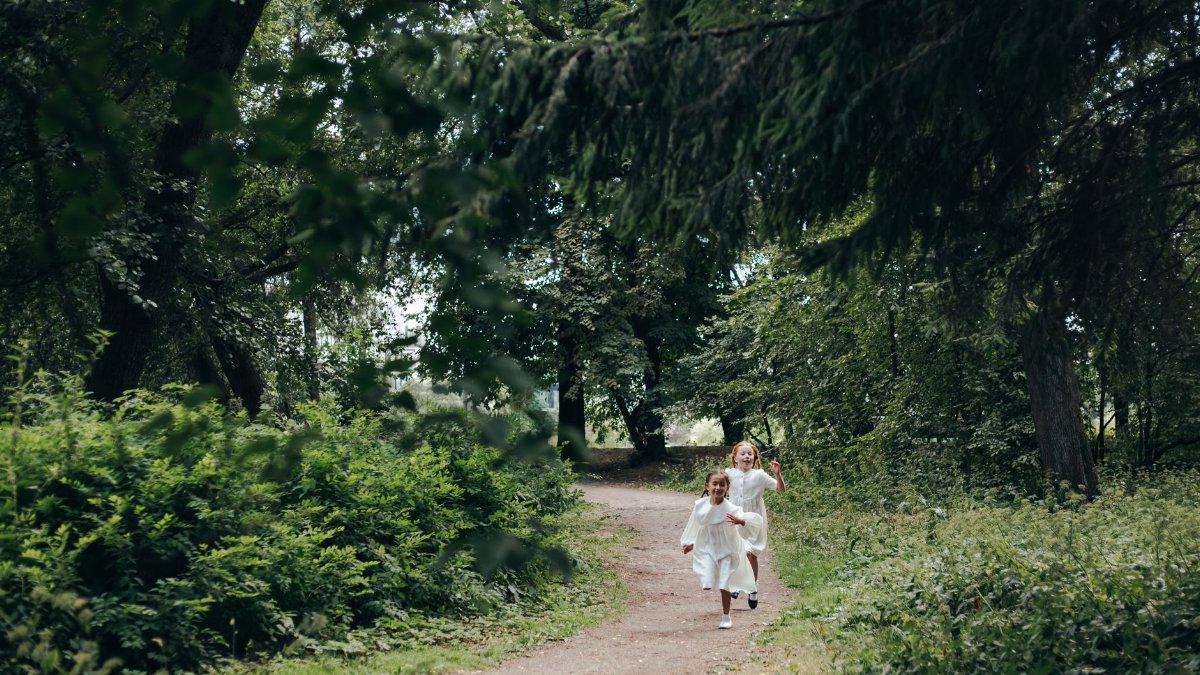 Two children in white dresses running on a forest path surrounded by lush greenery.