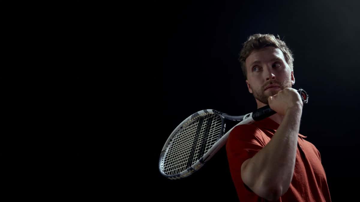 A thoughtful male tennis player holding a racket, set against a dark background.