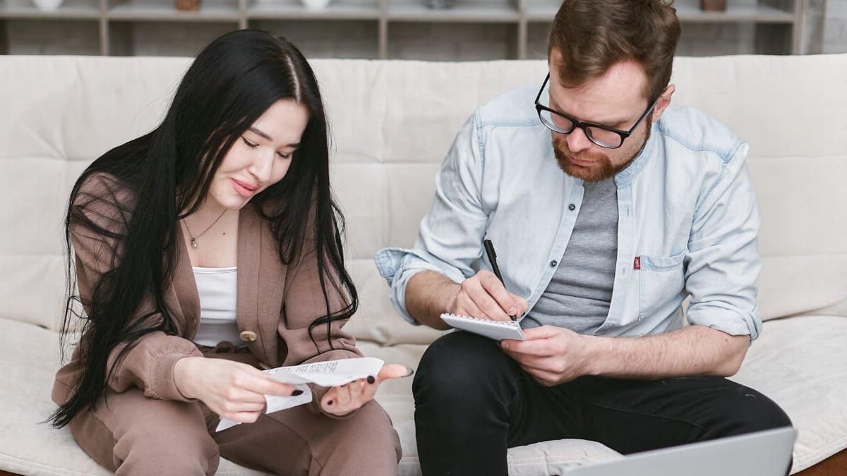 A man and woman sitting indoors, reviewing expenses with notebooks and money on the table.