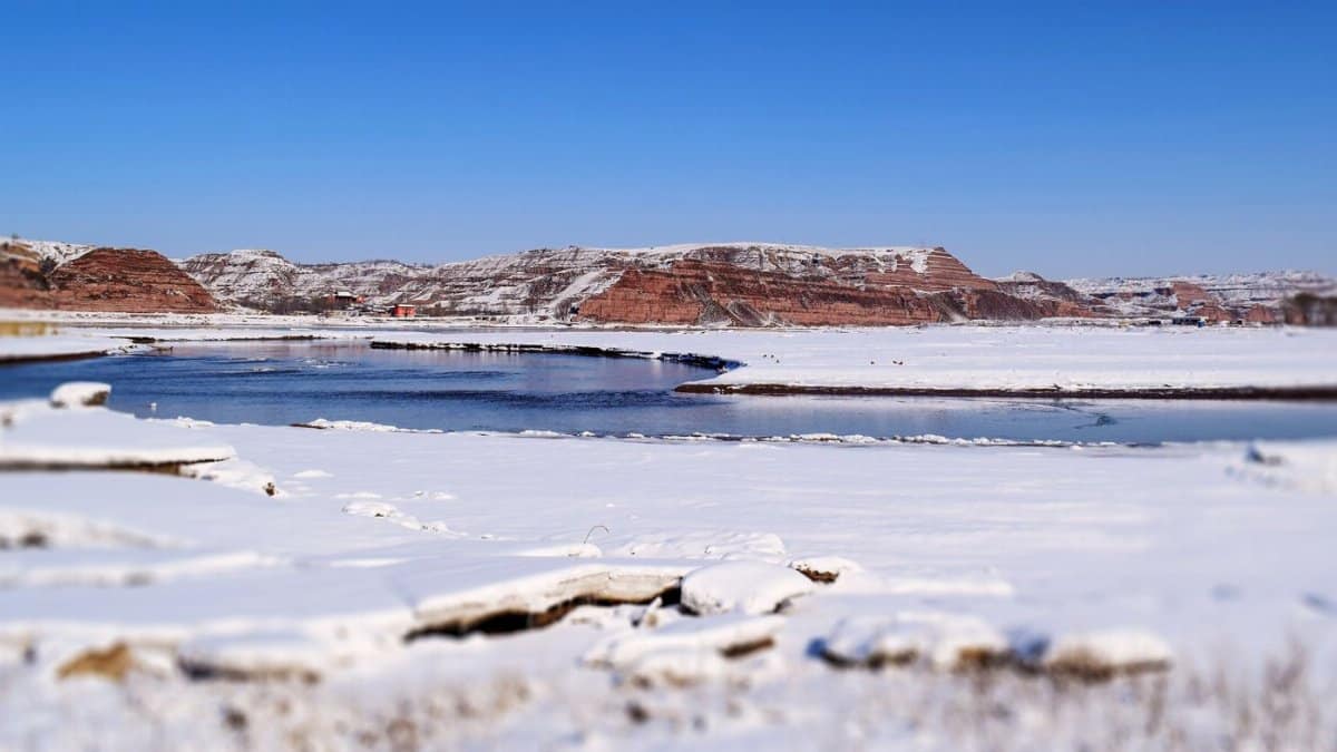 Winter scene of a snowy landscape with a river and red rock formations under a clear blue sky.