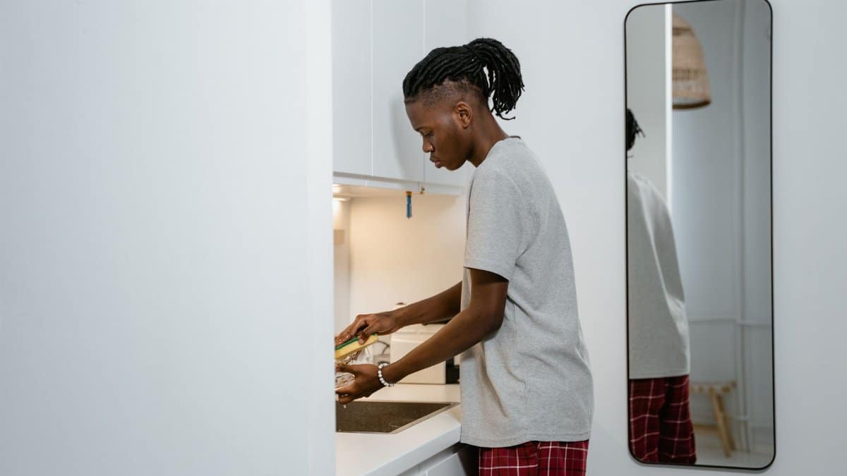 A young man in a gray t-shirt washing dishes in a modern kitchen with a mirror reflection.