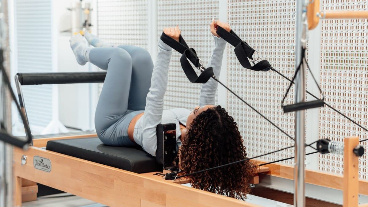 A woman wearing a gray tracksuit exercises on a Pilates reformer in a modern studio setting.
