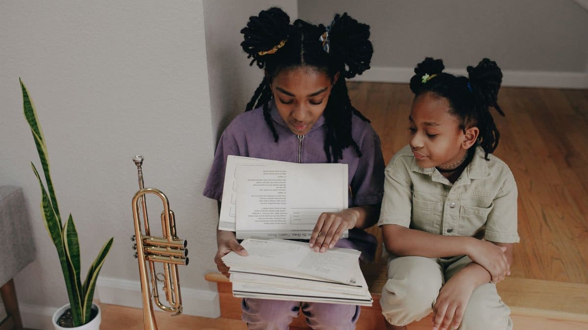 Two sisters reading music indoors with a trumpet nearby, focused and calm.