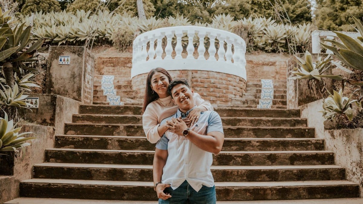 A smiling couple embraces on outdoor steps in a lush garden setting.