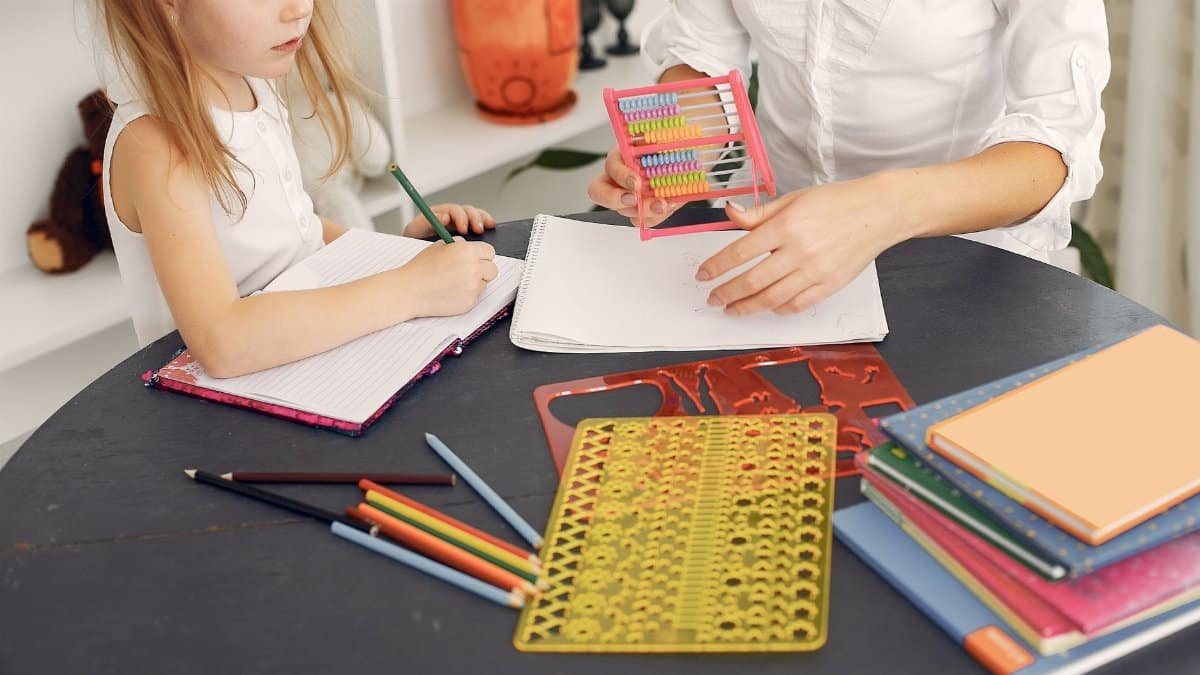 From above of little girl sitting at table with school items and textbooks while writing in notebook with pencil together with unrecognizable young home teacher in white shirt with abacus in hand during early home education