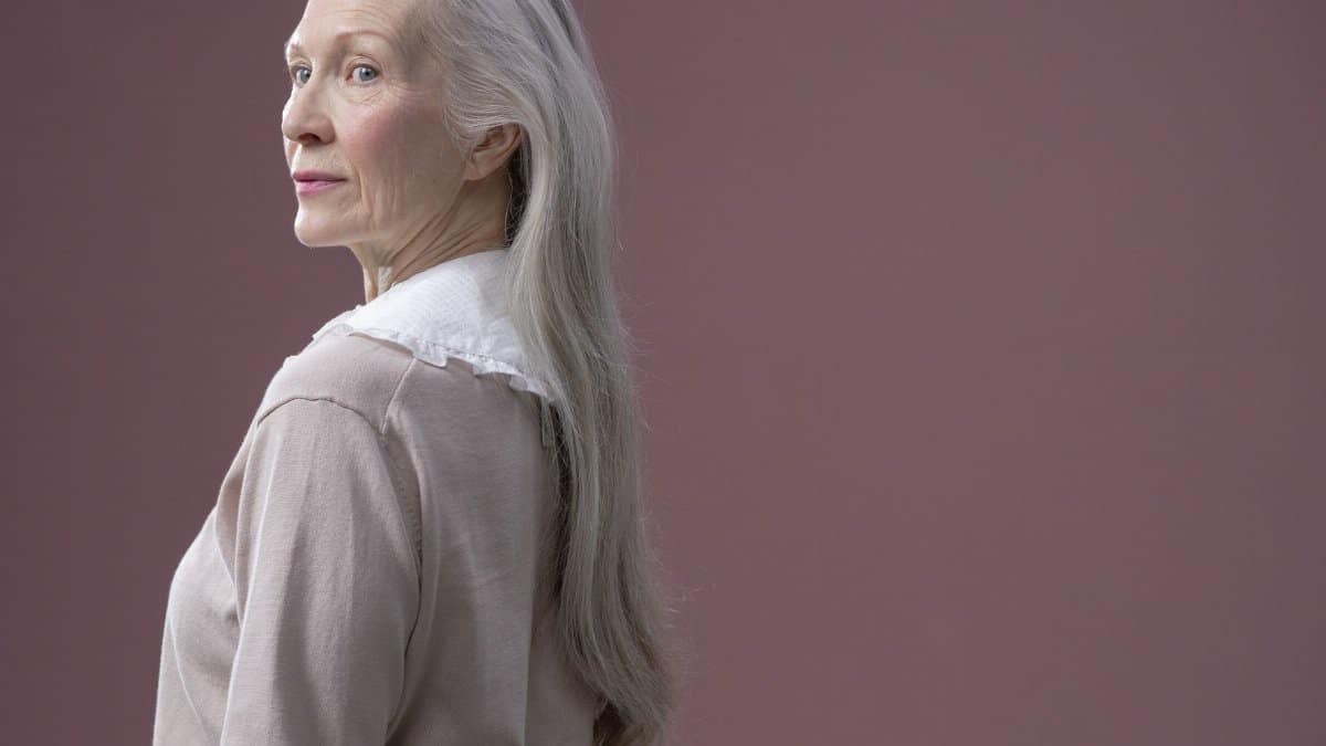 A sophisticated senior woman with long gray hair stands in profile against a solid background.