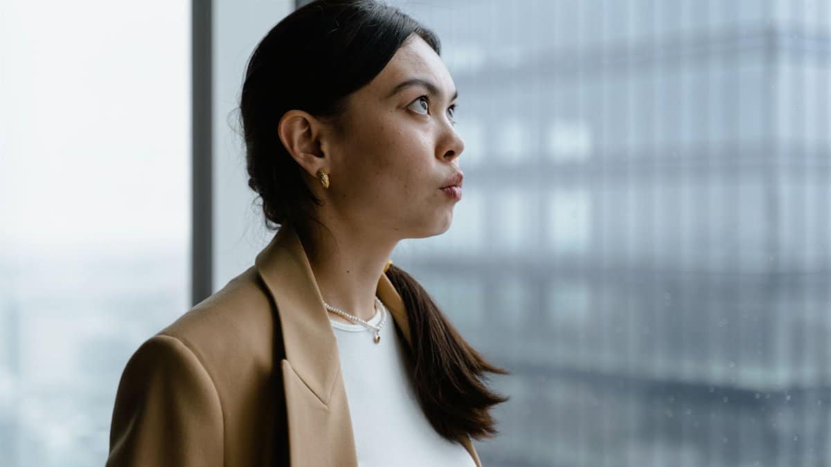 Profile of a woman in a brown blazer looking out of a window with urban background.