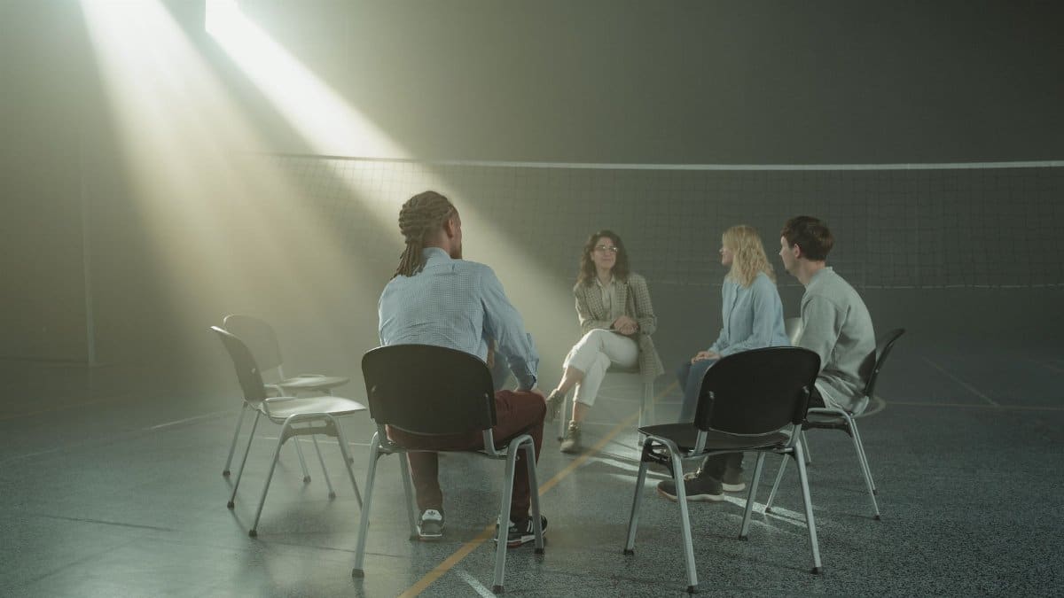 A group of diverse adults in a support meeting in a sunny indoor gym setting.