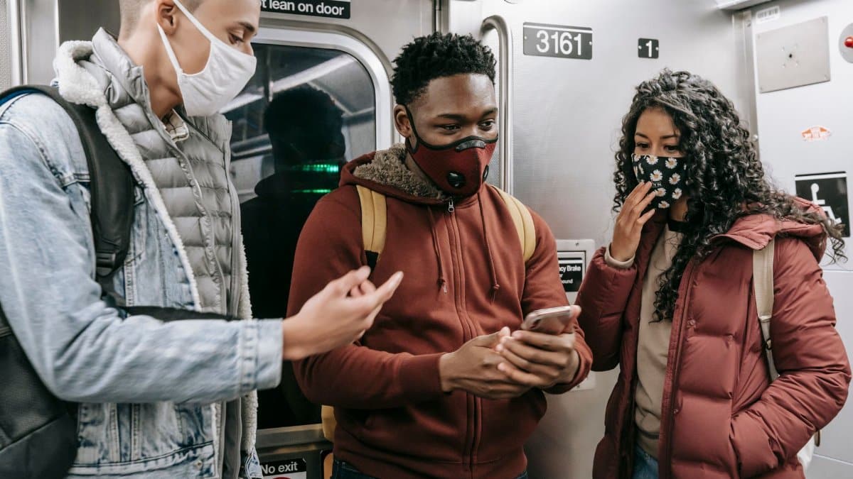 Group of anonymous young multiracial people in protective masks and warm clothes with backpacks talking while standing with cellphone in light train in underground