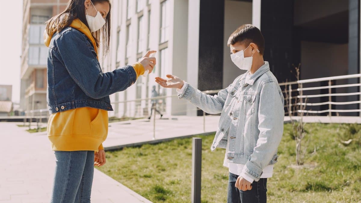 Side view of young boy and girl in casual wear and medical masks standing near modern building on street and disinfecting hands while using antibacterial hand sanitizer