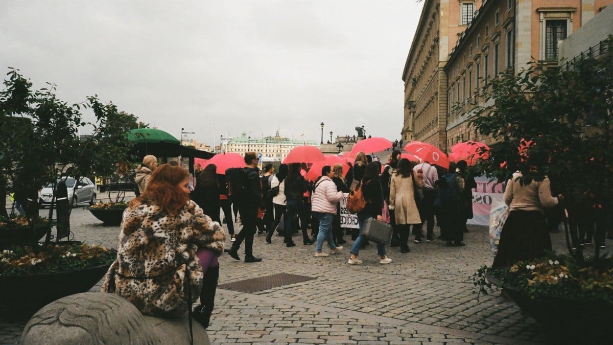 A lively group gathers outdoors under pink umbrellas, captured in a bustling urban setting.
