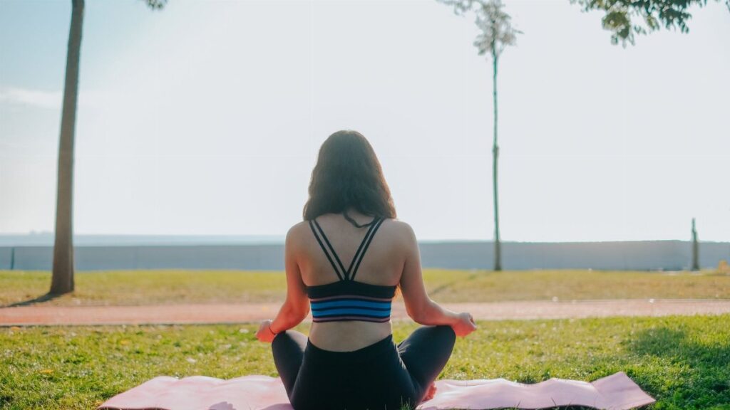 A woman practicing meditation on a yoga mat in a sunny park setting.