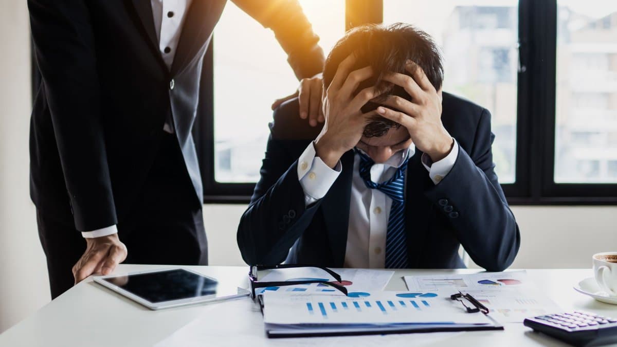 An overwhelmed businessman grasps his head, surrounded by reports in a bright office.