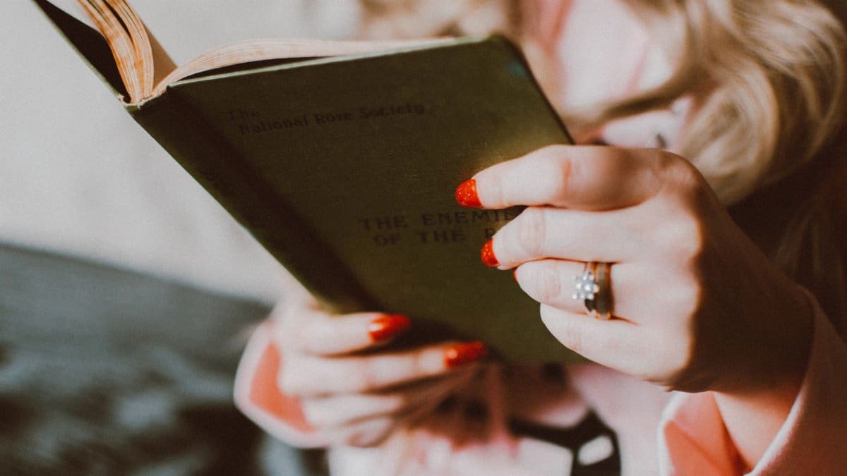 Close-up of a woman with red manicured nails and a ring holding a book indoors.