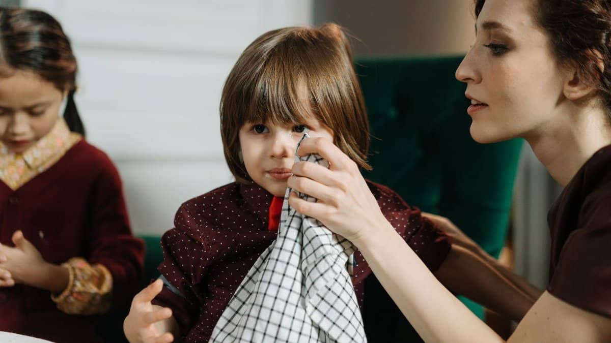 Woman assisting a child with a napkin during a meal indoors, highlighting care and family bonding.