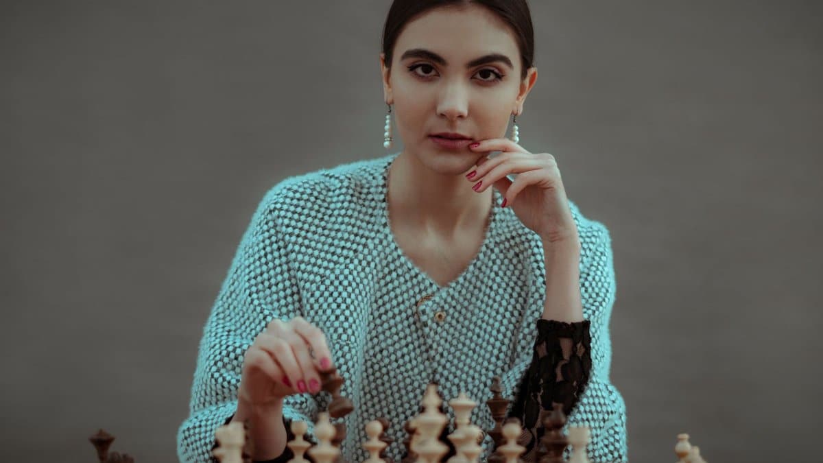Focused ethnic female player making chess move while playing game at table against gray background