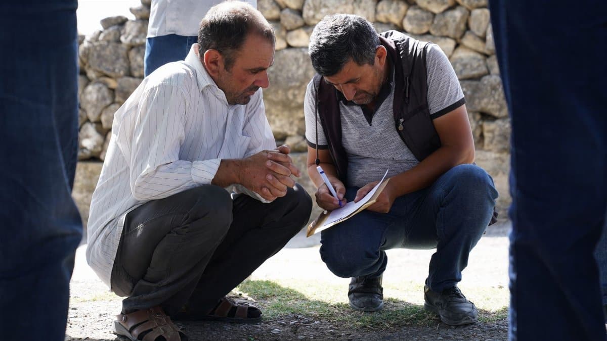 Two men squatting and analyzing paperwork outdoors in a sunny setting.