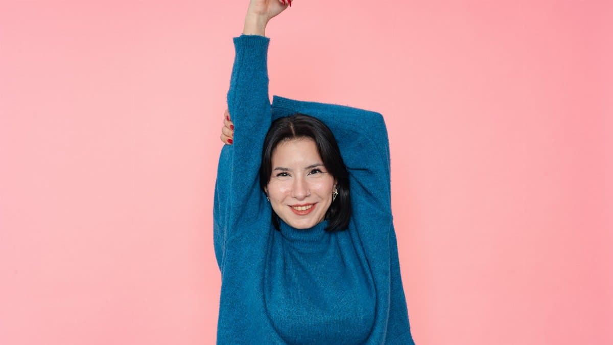 Cheerful woman stretching in a blue sweater with a pink backdrop, expressing joy and positivity.