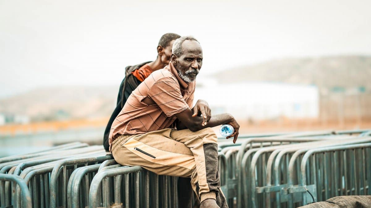 Two men sit on barriers in Tadjoura, Djibouti, with a serene background.