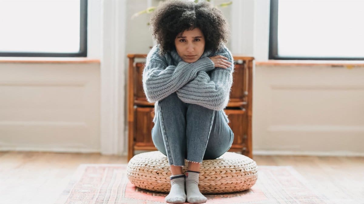 Full body of sad African American female looking at camera while sitting on rattan mat in light room at home