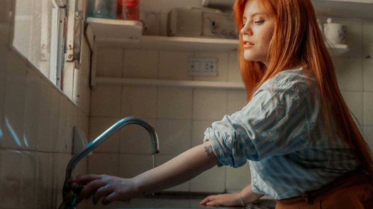 Portrait of a young woman in a kitchen adjusting a faucet with natural lighting.