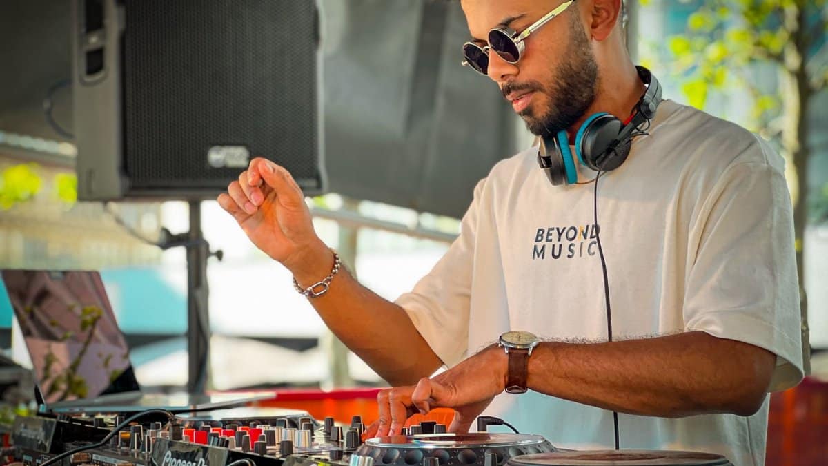 A stylish DJ wearing sunglasses and headphones mixes tracks outdoors using a turntable.