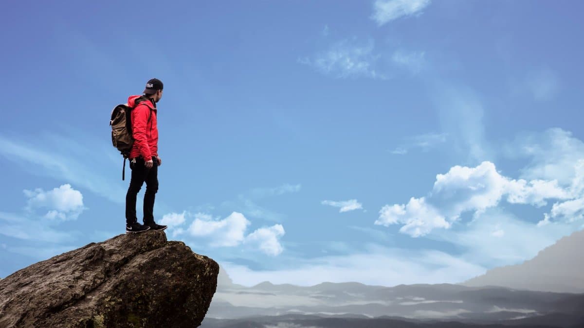 Hiker in red jacket stands on a cliff edge, overlooking a scenic mountain view under a bright blue sky.