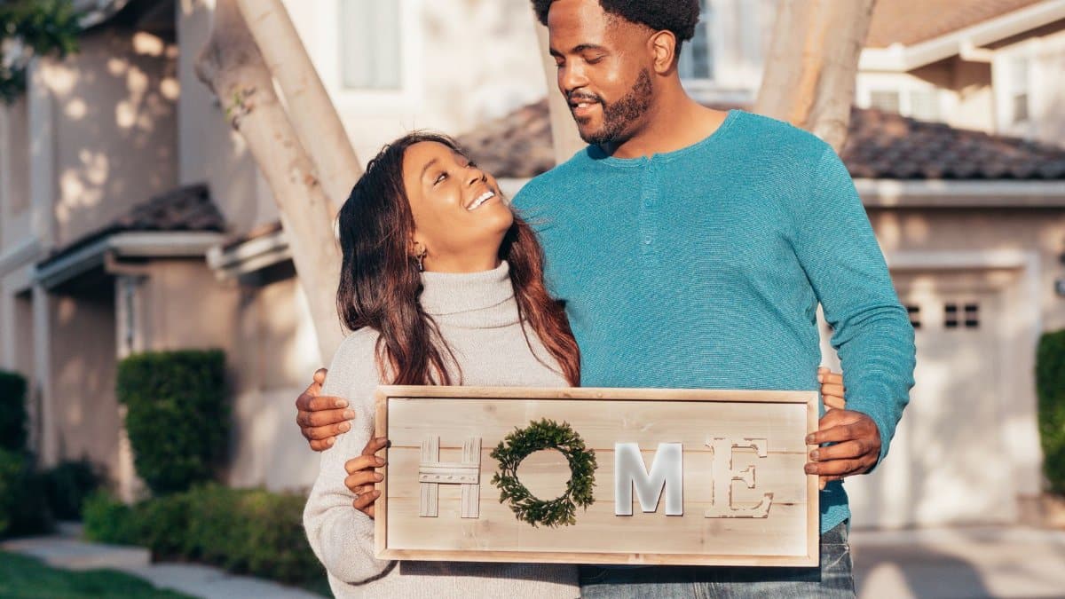 Joyful couple celebrating home purchase, holding 'Home' sign outdoors.