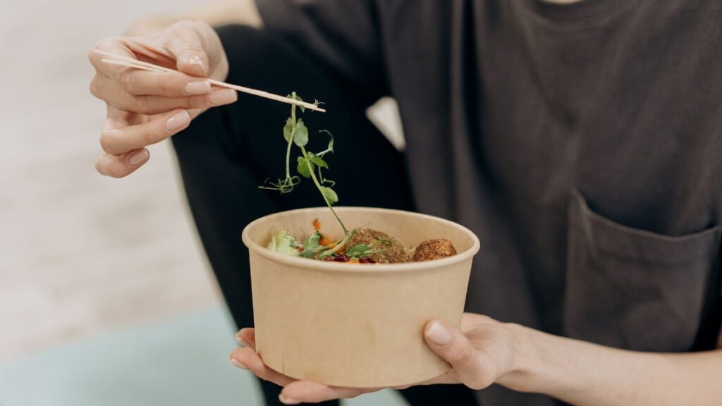 A woman holding a paper bowl filled with a healthy vegetarian meal in an indoor setting, promoting wellness.
