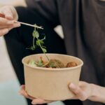 A woman holding a paper bowl filled with a healthy vegetarian meal in an indoor setting, promoting wellness.