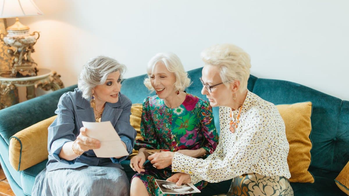 Three senior women joyfully reminiscing over old photos on a cozy couch.
