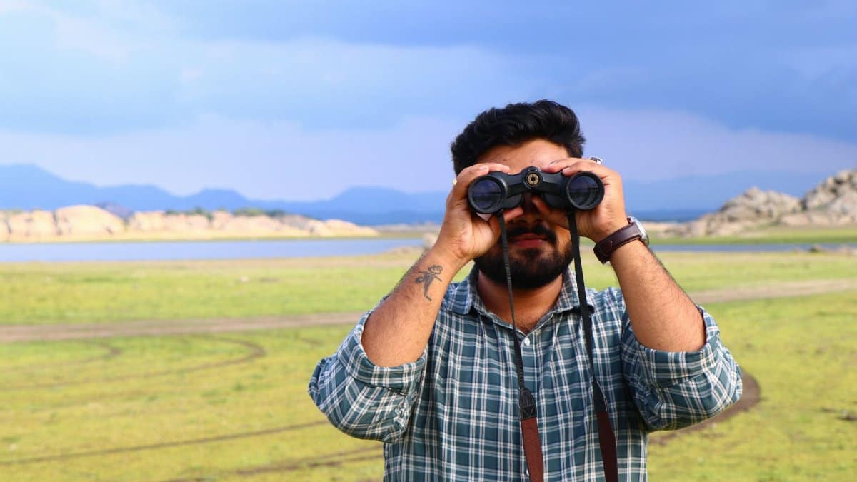 Man with beard and tattoo using binoculars in a grassy field under a blue sky.