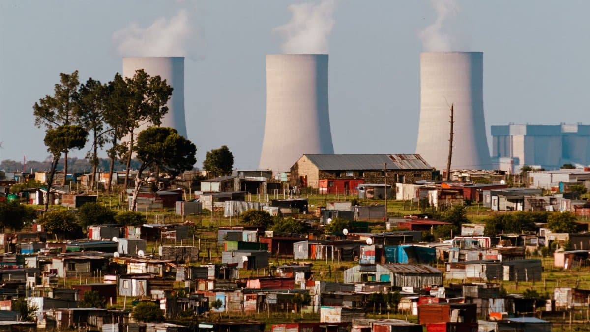 Cooling towers emit smoke over a vibrant South African settlement, highlighting industrial impact.