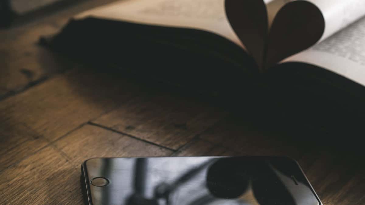 A smartphone rests on a wooden table beside an open book with pages forming a heart.