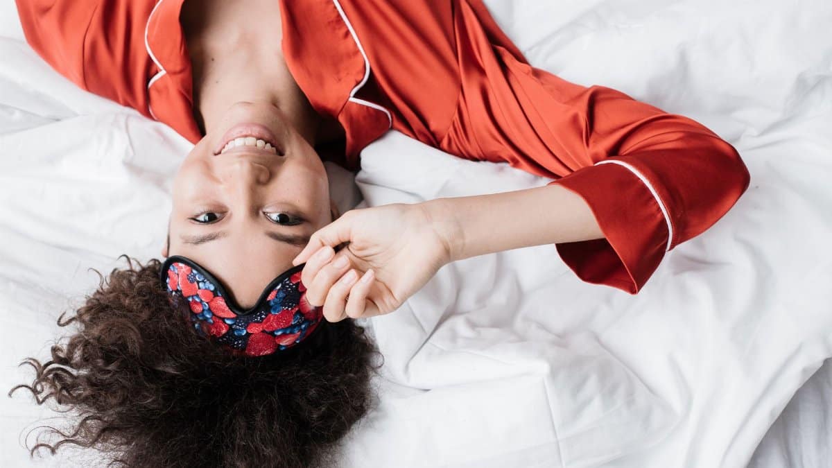 Happy woman in red sleepwear smiling while adjusting her sleep mask. Top view.