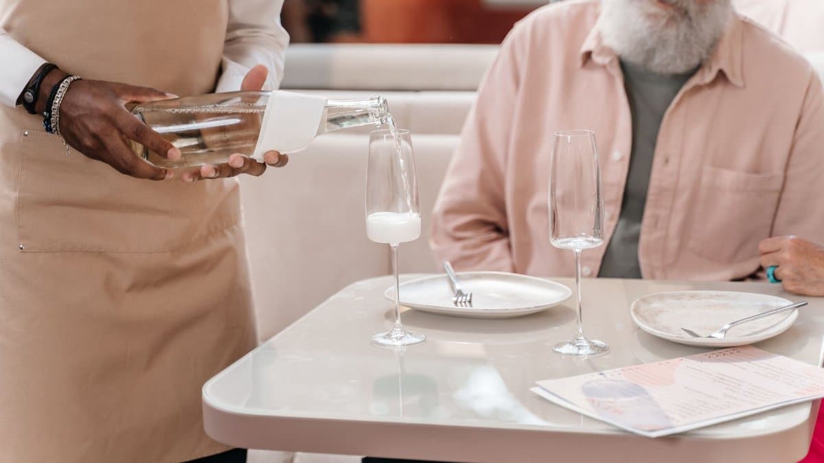 A waiter pouring a drink for an elderly man seated at a restaurant table. Focus on hospitality.