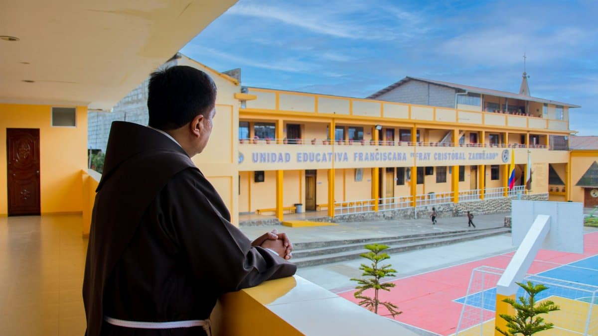 Franciscan monk overlooks educational institute from balcony, highlighting religious presence.