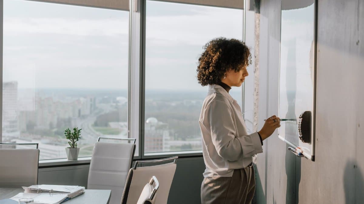 Confident woman with curly hair writes on a whiteboard in a modern office with city views.