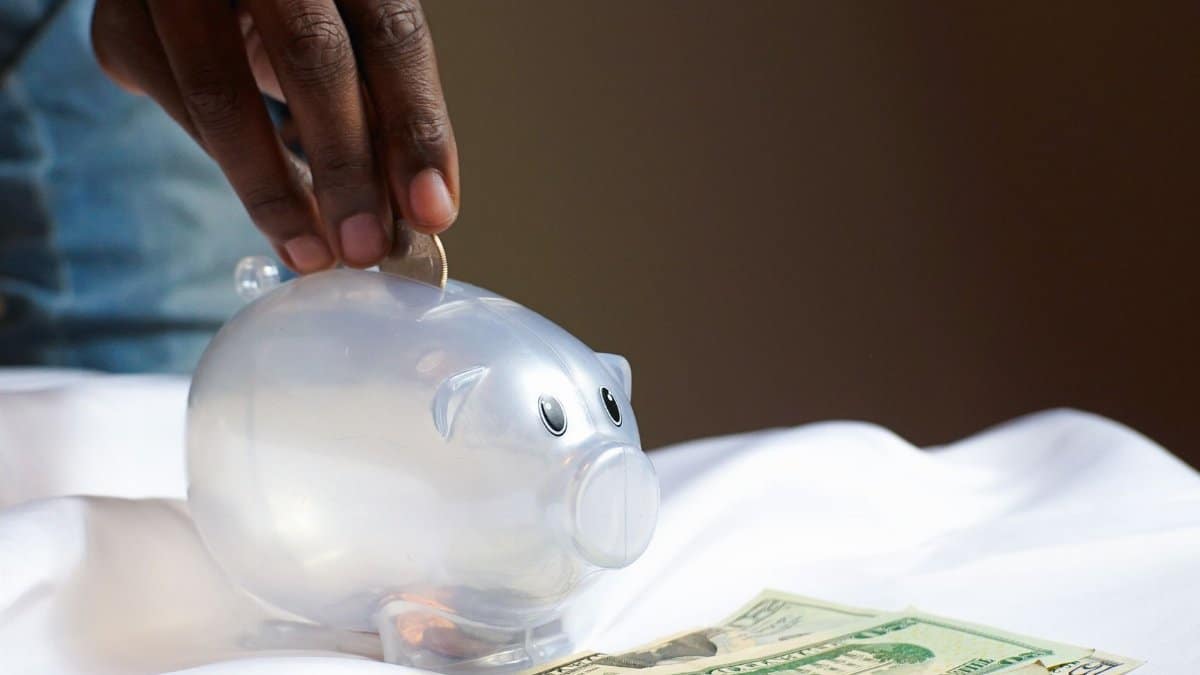 A hand placing a coin in a piggy bank with dollar bills nearby signifying savings and finance.